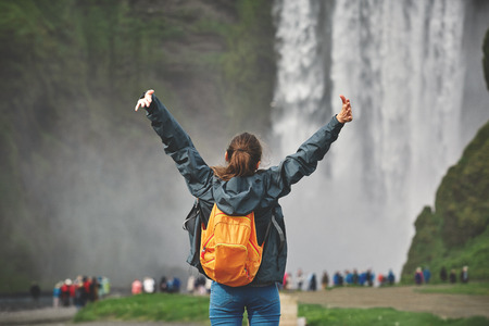 Girl in waterproof clothing stands on background of Skogafoss waterfall in Iceland. back view, woman holds hands upの写真素材