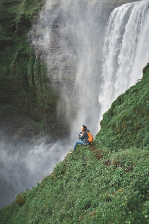 Girl in waterproof clothing sits on the cliff on background of Skogafoss waterfall in Iceland.の写真素材