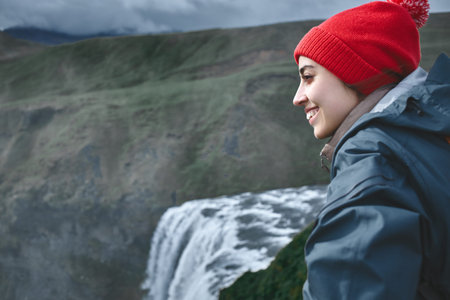 Girl in waterproof clothing stands on the cliff on background of Skogafoss waterfall in Iceland. View from aboveの写真素材