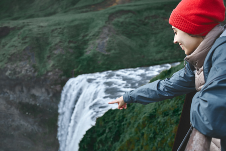 Girl in waterproof clothing stands on the cliff on background of Skogafoss waterfall in Iceland. View from aboveの写真素材