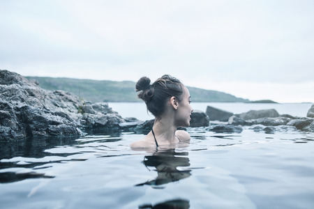 Young cheerful girl swimming in water of pool looking away on background of sea, Iceland, West Fjords. back view, Woman washing and covering her face with her handsの写真素材