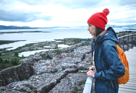 Girl in warm clothing observing surroundings on background of lake and mountains of Iceland.