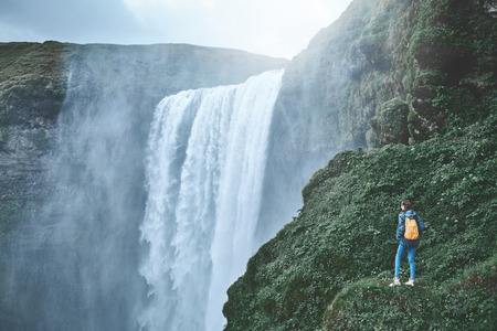 Girl in waterproof clothing stands on the cliff on background of Skogafoss waterfall in Iceland. back view, woman holds hands upの写真素材