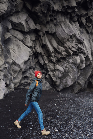 woman traveler with small orange backpack walking on Kirkjufjara black sand beach, southern Iceland.の写真素材