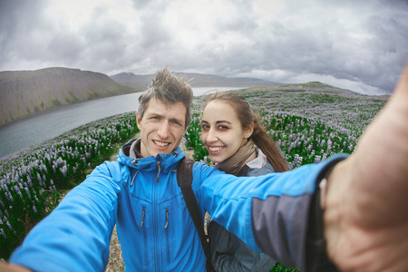 Couple in warm outdoor clothing makes selfie photo on background of mountains and sea of Iceland. A field of flowering lupines in the background. Beautiful summer landscape in Icelandの写真素材