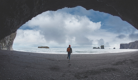 woman traveler with small orange backpack walking on Kirkjufjara black sand beach, southern Iceland.の写真素材