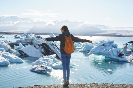 back view of girl in warm clothing with orange backpack in Ice Lagoon in Iceland with background of sea and sky.の写真素材
