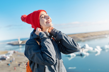 portrait of happy young smiling woman in warm clothing and knitted red hat with orange backpack in Ice Lagoon in Iceland with background of sea and sky. a woman is wearing a hat with a happy look and a smileの写真素材