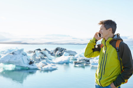 portrait of man in waterproof jacket with orange backpack using smartphone in Ice Lagoon in Iceland...