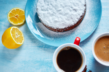 whole biscuit sprinkled with powdered sugar on the blue plate and two red cups of coffee, cut lemon and badon on blue wooden backgroundの写真素材