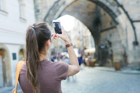 a young smiling woman tourist in sportswear walking in the center of Prague with a phone and taking photo and selfie. travel guide, tourism in Europe, woman tourist with smartphone on the streetの写真素材