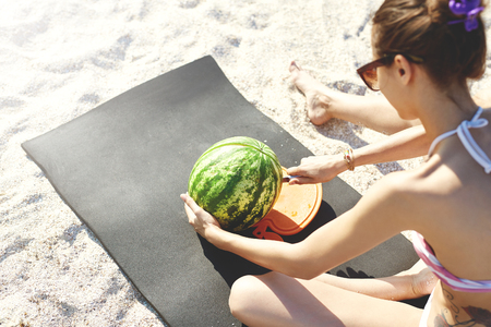 young beautiful woman is going to cut and eat watermelon on the beach at hot summer day, back view.の写真素材