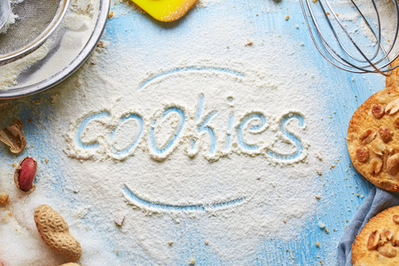 word Cookie written on flour and top view of product set for cooking cookies, kitchenware and several round cookies with peanuts on the blue wooden table. word Cookiesの写真素材