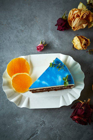 composition of piece of cutted mousse cake with blue glaze on a white paper dish, sliced oranges and dried roses on a dark gray concrete background. Top view with copyspace