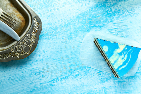 piece of mousse cake with blue glaze on the white-blue wooden background. Looking like silver, vintage metal dish, fork, knife on the table. Horizontal Top view with copyspace