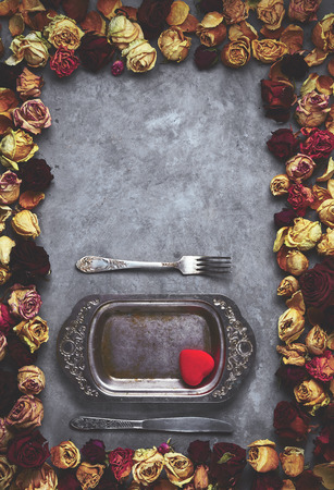 knife, fork and vintage metal dish with small red heart in frame from dried roses buds on the gray concrete background. Valentines Day holiday concept, top view with copyspaceの写真素材