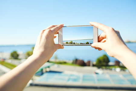 first person view of woman photographing a cityscape with river side on mobile phoneの写真素材