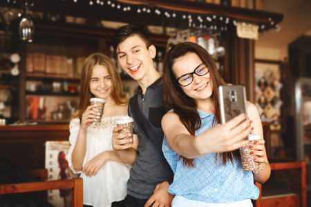 Group of friends enjoying in cafe together. Young people meeting in a cafe. Young men and women with paper cups standing at bar counter in cafe and making selfieの写真素材