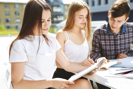 the guy student explains to the girls something and shows in a notebook. Portrait of three happy high school students. students friends learn, do homework together, spend time and have fun.の写真素材