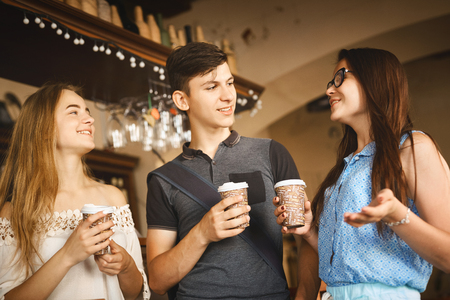 Young man and women with paper cups standing at bar counter in cafe, laughing and talking. Group of friends enjoying in cafe together. Young people meeting in a cafe.の写真素材