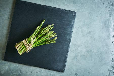 bunch of fresh raw garden asparagus on rectangular rough black slate board on gray concrete background. Ingredients for cooking. Food background. Top view with copy space.の写真素材