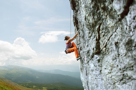 side view of young slim woman rock climber in bright orange pants climbing on the cliff against a blue sky. girl climbs on a vertical flat rocky wall and making hard move. Copy spaceの写真素材