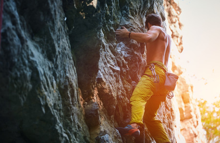 muscular man rock climber in bright yellow pants climbing the challenging route on the cliff. strong bearded rock climber climbs on a rocky wall, bottom viewの写真素材