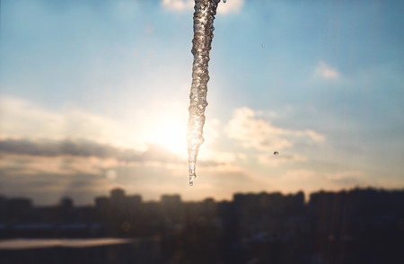 big long icicle against a sunset sky in the city. icicle hanging from the roof of the house, the view from the windowの写真素材