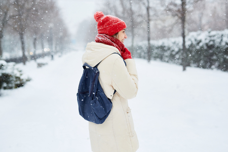 Cheerful smiling woman in white down jacket and red cap, scarf and mittens walking on the snowy street after blizzard in city. winter city after blizzards and snowfall.の写真素材