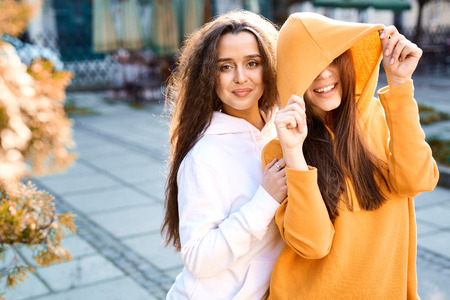 two young girls walking city having fun. joyful Women in bright colored hoodies walking, laughing and posing on the street, pulling the hoods over the headsの写真素材