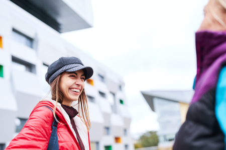 Portrait of a young smiling woman in red jacket,gray scarf and cap. Woman walking, talking and laughing outdoors on the street with girlfriend at sunny autumn or spring dayの写真素材