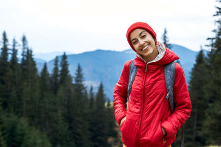 woman hiker with backpack, wearing in red jacket and cap, standing on the mountains and woods background, Western Ukraine.の写真素材