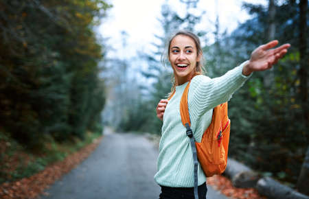 beautiful smiling woman hiker with small backpack standing on the road in the forest. Woman gives a hand to someone, Follow me concept.の写真素材