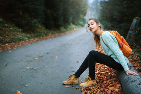 girl traveler in yellow boots and mint sweater, with small orange backpack, sitting on the big log near road in autumn forestの写真素材
