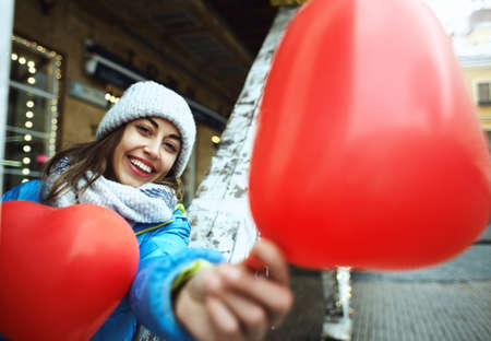 happy smiling young woman in warm winter knitted cap and scarf is standing outdoors with a heart shaped red balloonsの写真素材