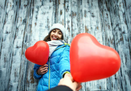 horizontal image of a happy smiling young woman is standing outdoors with a heart shaped red balloons on the old wooden door backround. woman is accepting a balloon from someoneの写真素材