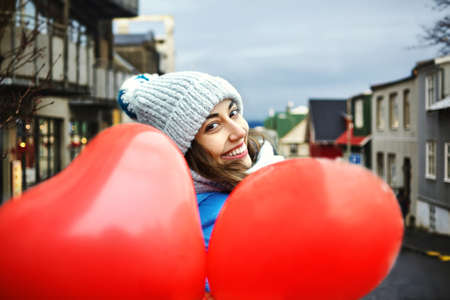 happy smiling young woman in warm winter knitted cap and scarf is walking outdoors with a heart shaped red balloonsの写真素材