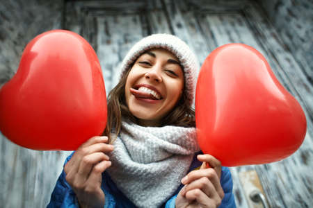 horizontal outdoors image of a happy smiling young woman in warm winter knitted cap and scarf with a heart shaped red balloons. woman shows a funny face sticking her tongue outの写真素材