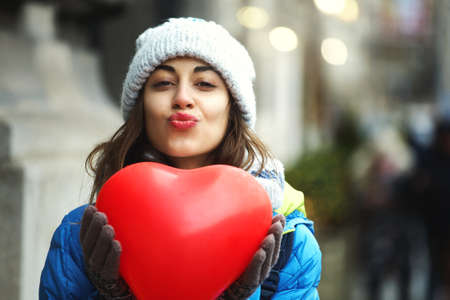 horizontal image of a cheerful young woman with a heart shaped red balloons. woman blows kiss, demonstrates her good feelings, says Love you on distance. Young pretty female model makes air kissの写真素材