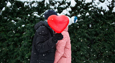 happy hugging and kissing couple in warm winter clothes is standing outdoors with a heart shaped red balloon on the green fence backgroundの写真素材