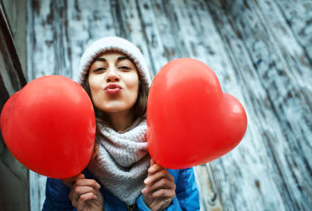 horizontal image of a cheerful young woman with a heart shaped red balloons. woman blows kiss, demonstrates her good feelings, says Love you on distance. Young pretty female model makes air kissの写真素材