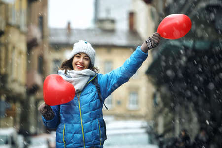 happy smiling young woman in blue jacket and warm knitted cap and scarf is standing outdoors on the snowy street holding a heart shaped red balloons in hands.の写真素材