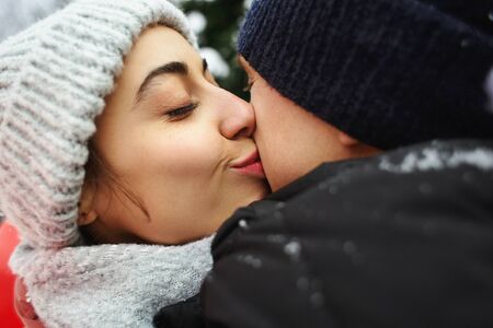 happy hugging and kissing couple in warm winter clothes is standing outdoors with a heart shaped red balloon on the green fence backgroundの写真素材