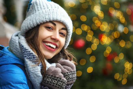closeup horizontal image of a side looking happy smiling woman in warm winter clothes against the background of bright Christmas lightsの写真素材