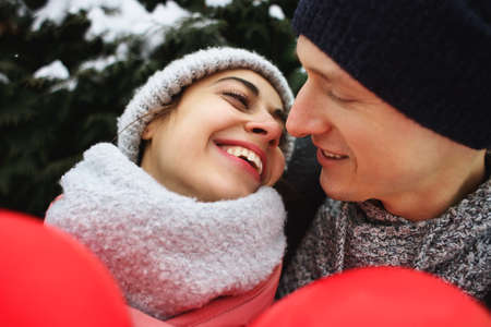 happy hugging and kissing couple in warm winter clothes is standing outdoors with a heart shaped red balloon on the green fence backgroundの写真素材