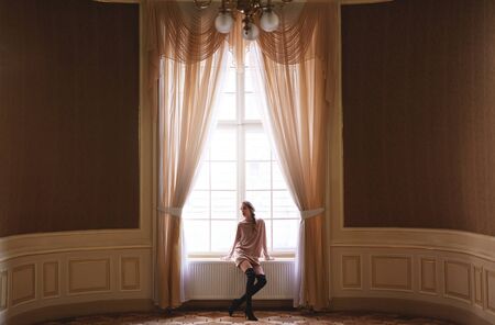 pretty elegant woman sitting at the big window and posing in the big wide palace hallの写真素材
