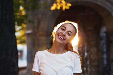 portrait of a young attractive smiling woman in white t-shirt standing on the street in a sunset backlightの写真素材