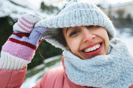 happy joyful smiling woman in light knitted cap and scarf walking outdoors at frosty snowy winter day. a woman puts on a warm hat