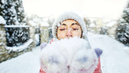 cute happy joyful smiling woman in warm winter clothes holding a snow in a both palms and blowing snow to the cameraの写真素材