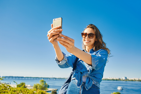 young smiling attractive woman in jeans clothes makes selfie or photographing something on smartphone at sunny day on the blue sky background. woman posing in city scape.の写真素材